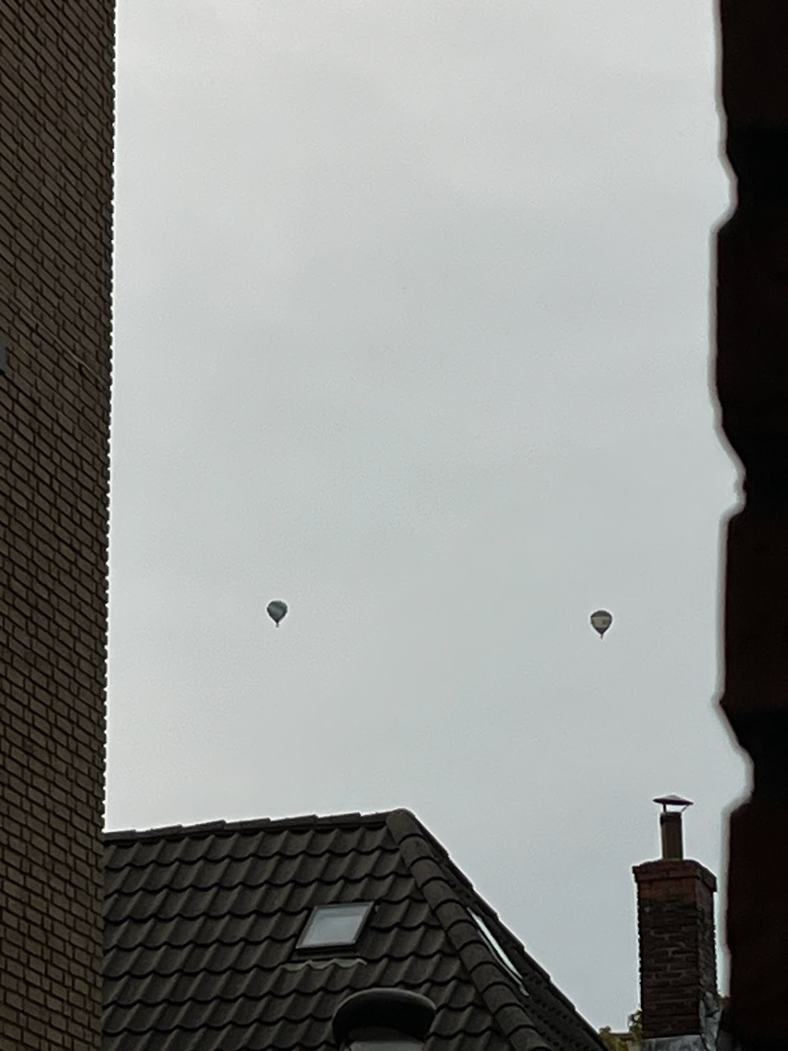 A photograph of two hot air balloons in the sky. They're framed by walls and a nearby roof.