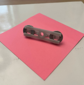 Pictured, from the back and front, laying on a pink post-it note: A small, plastic thingamabob, of, in the front, two horizontal bars connected by a circular hole, and in the front, one horizontal bar through which the aforementioned circular hole goes. On the back it's also sort of a gear thing? It's holding two small metal wheels, keeping them in place with the little speedbumps that are on the back horizontal bar. Image descriptions are hard.