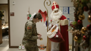 Screencap. Inside the base, Jan is dressed like Sinterklaas, holding a bag of oranges. There's Christmas decorations everywhere, and clogs set against the wall. A young soldier is about to walk away from him.